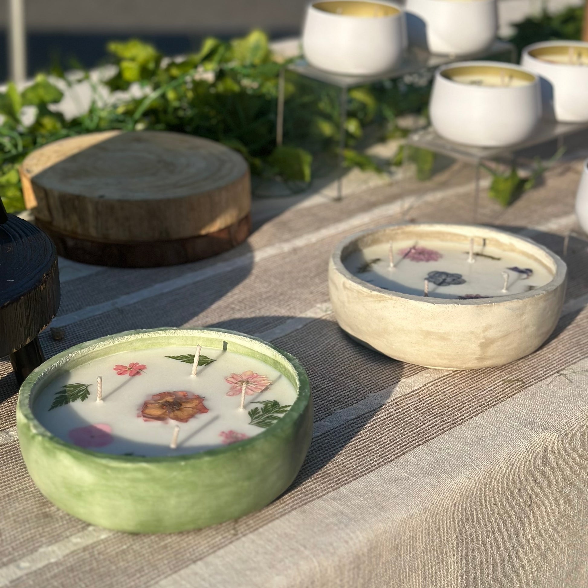 Green and Cream Concrete Bowls 12 ounce Containers with Dried Pressed Perennial Flowers on Display Table. More Small White Metal Containers in the Background on a Cream and Brown Striped Table Cloth and Green Vine and Wooden Slabs as Decoration  