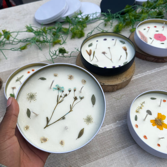 One White Container 12 Ounce Candle with Blue and White Dried Pressed Flowers in Hand and Black and White Containers Behind on Display Table
